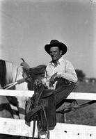 [Unidentified cowboy leaning on saddle which is draped over fence]