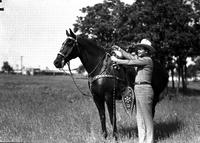[J. Wills standing next to dark horse wearing silver saddle. Trees in background]