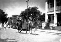 [Two unidentified older cowboys on horses riding down residential street]