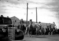 [Pairs of mounted riders, both cowboys & cowgirls, parading down street]