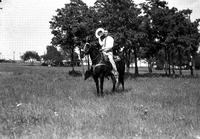 [J. Wills with hat in hand on dark horse wearing silver saddle. Trees in background]