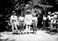 [Gene Autry standing with three cowgirls at park; picnic tables and other rodeo people behind them]