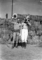 [Gene Autry & unidentified man with hats raised over heads in front of hay bales]