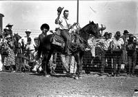 [Unidentified Cowboy on horseback doffing hat and holding rope; onlookers behind wire fence]