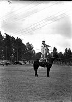 [Unidentified Cowgirl standing atop stationary horse spinning large rope loop about herself & horse]