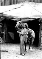 [Unidentified cowgirl standing beside horse in front of tent]