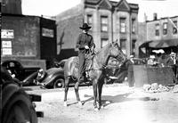 [Unidentified cowboy on horseback with rope in hand; people milling about in background]