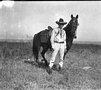 [Possibly Jack Hughes in light colored outfit, big hat standing in field with horse]