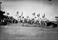 [Line of one cowboy & four cowgirls bearing flags moving down city street]