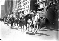 [Two unidentified cowgirls atop horses leading a parade followed by several cowboys]