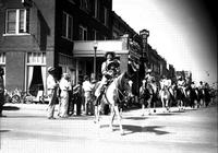 [Unidentified Cowgirl on horse with two western-attired children in her lap leading line of riders]