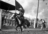 [Unidentified Cowboy on horseback carrying flag "Swift's Jewel Cowboys" galloping past grandstand]