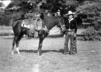 [Possibly Silver Dollar Brady  in chaps, gloves, neckerchief, and tall hat standing with horse]