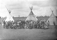 [Group of Indians and white men, &  women in formal attire gathered in front of tipis]