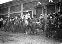 [Line of standing and mounted cowboys and one clown in front of building]
