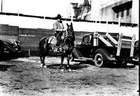 [Unidentified Cowboy on horseback between truck and car holding rope loop in hand]