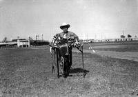 [J. Wills standing with Silver mounted saddle, and bridle, in grassy field, stadium in background]