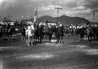 [Unidentified Cowboy on horse with a cowgirl on horseback either side; line of rodeo people behind]