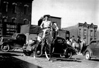 [Possibly Cy Compton on horse with cars, trucks, additional cowboys, and buildings in background]