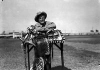 [Unidentified Cowboy leaning on Wills' silver mounted saddle in grassy field, stadium in background]