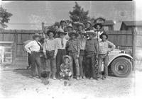 [Group of unidentified cowboys standing on & in front of car; Midget clown squatting in front]