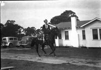 [Hatless unidentified cowgirl atop trotting horse; Bungalow and autos in background]