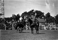 [Unidentified Cowboy standing atop stationary horse throwing a rope loop in front of passing rider]