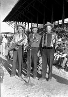 [Three unidentified men in western wear standing behind microphone with musical instruments]