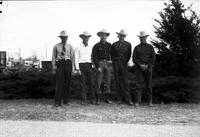 [Five unidentified cowboys standing in line in front of low shrubbery]