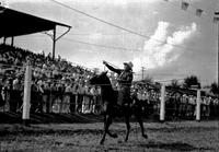 [Unidentified cowboy spinning a rope loop around himself on moving horse in front of grandstand]