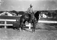 [Unidentified Cowboy on horseback with stable/barn in background]