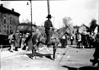 [Unidentified Cowboy on horseback with rope in hand; people milling about in background]