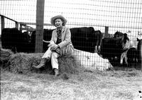 [Older cowgirl sitting on hay bale in front of livestock fence]
