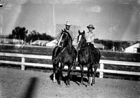 [Two unidentified cowboys on horseback with stable/barn in background]