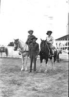 [Unidentified cowboy standing between two cowgirls on horses]