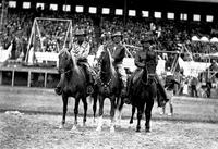 [Leo Cremer with two unidentified cowboys on horseback in front of grandstand]