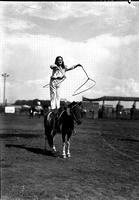 [Young unidentified Cowgirl standing behind saddle on pony and spinning a rope with each hand]
