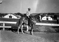 [Unidentified young Cowboy on horseback with stable/barn in background]