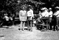 [Gene Autry and Leo Cremer standing side by side in park surrounded by rodeo people @ picnic tables]