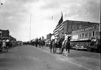 [Young Cowboy on horseback bearing American Flag is followed other riders on downtown street]