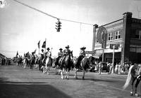 [Two unidentified cowgirls on horseback crossing intersection followed by riders]