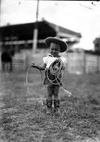 [Very young cowgirl in boots and shorts holding coil of rope with left hand and rope loop in other]