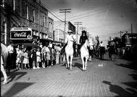 [Two unidentified cowgirls on horseback followed by other riders in front of corner drugstore]