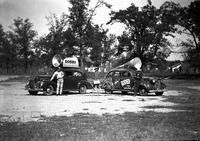 [Two rodeo publicity cars each with megaphones on roof; three cowboys standing by them]