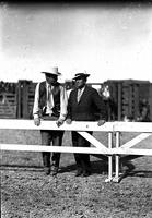 [Two unidentified men leaning on hurdle one in western wear the other in business attire]