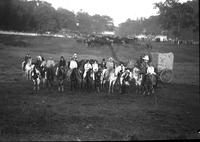 [Group of mounted cowboys and cowgirls with covered wagon behind them and horses in far distance]