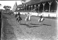 [Mounted Indian carrying American Flag, Cowboy and young cowboy side by side in front of stands]