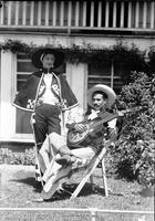[Possibly Jack Wolfe with guitar sitting in chair in chaps & Marie Wolfe stands by him in cap & hat]