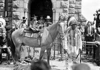 [Indian man in full feathered headdress standing by and holding reins of saddled horse]