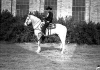 [J. Wills on White Horse wearing Silver mounted saddle. Bushes and Brick Building with tall windows]